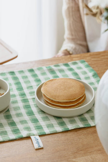 Three plain pancakes sit on a Soft Spot Soft Suction Plate, displayed atop a green and white checkered cloth on a wooden table. A bowl and an arm in the background highlight the convenience of Soft Spot’s food-grade silicone plate.