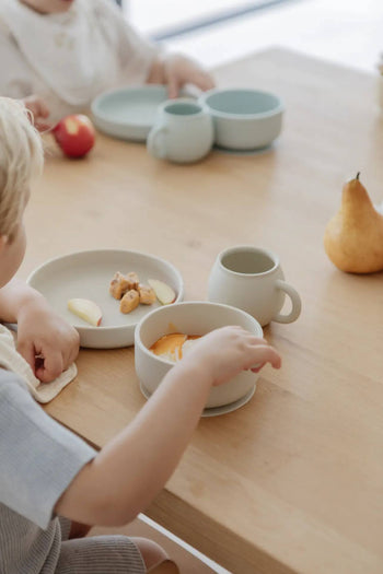 Two young children sit at a light wooden table with Soft Spot Soft Suction Plates, cups, and bowls in pale blue. One enjoys sliced fruit and snacks; a pear and apple rest nearby. The minimal scene feels safe and perfect for little ones.
