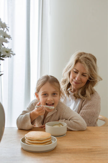 A smiling woman sits with a young girl, who eats from the Soft Spot Soft Suction Plate by Soft Spot. Pancakes are stacked nearby, and the plate’s strong suction grip helps keep mealtime easy in the softly lit, happy room.