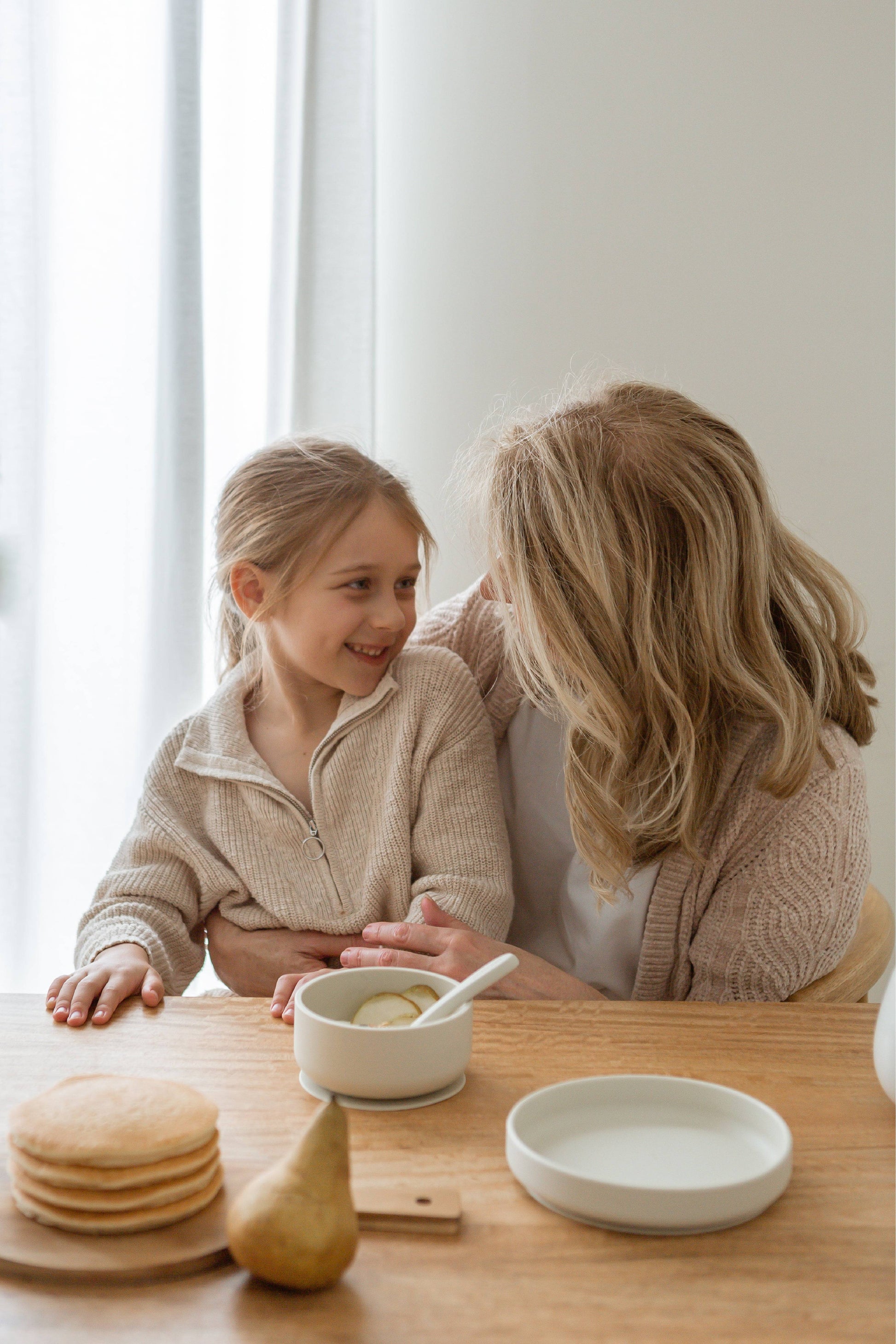 A woman and a young girl sit at a wooden table, smiling at each other. On the table are Soft Spot Soft Suction Plates by Soft Spot, ideal for kids, along with a bowl, spoon, pancakes, and a pear. Daylight filters through sheer curtains in the background.