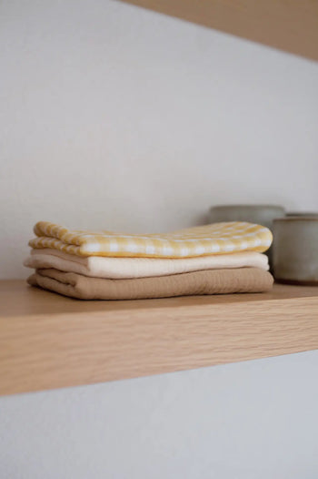 A stack of Soft Spot Soft Tea Towels (Set of 3) in beige, white, and yellow patterns rests on a wooden shelf beside two small ceramic containers.