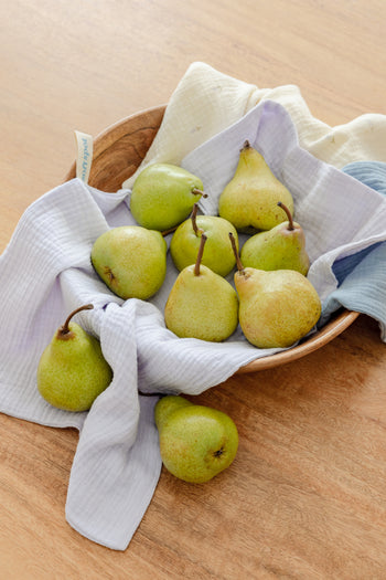 The Soft Spot Soft Tea Towels (Set of 3) by Soft Spot, in cream, blue, and white, hang by labeled loops from a wooden rack against a light gray wall—ideal for use as hand or tea towels.