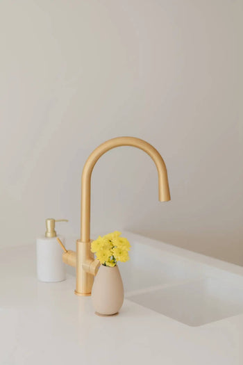 A minimalist kitchen sink with a gold faucet, the Soft Spot Soft Vase by Soft Spot holding yellow flowers, and a white soap dispenser on a clean white countertop.