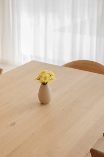 The Soft Spot Soft Vase with yellow flowers sits on a light wooden table, while sheer curtains let natural light brighten the minimalist space.
