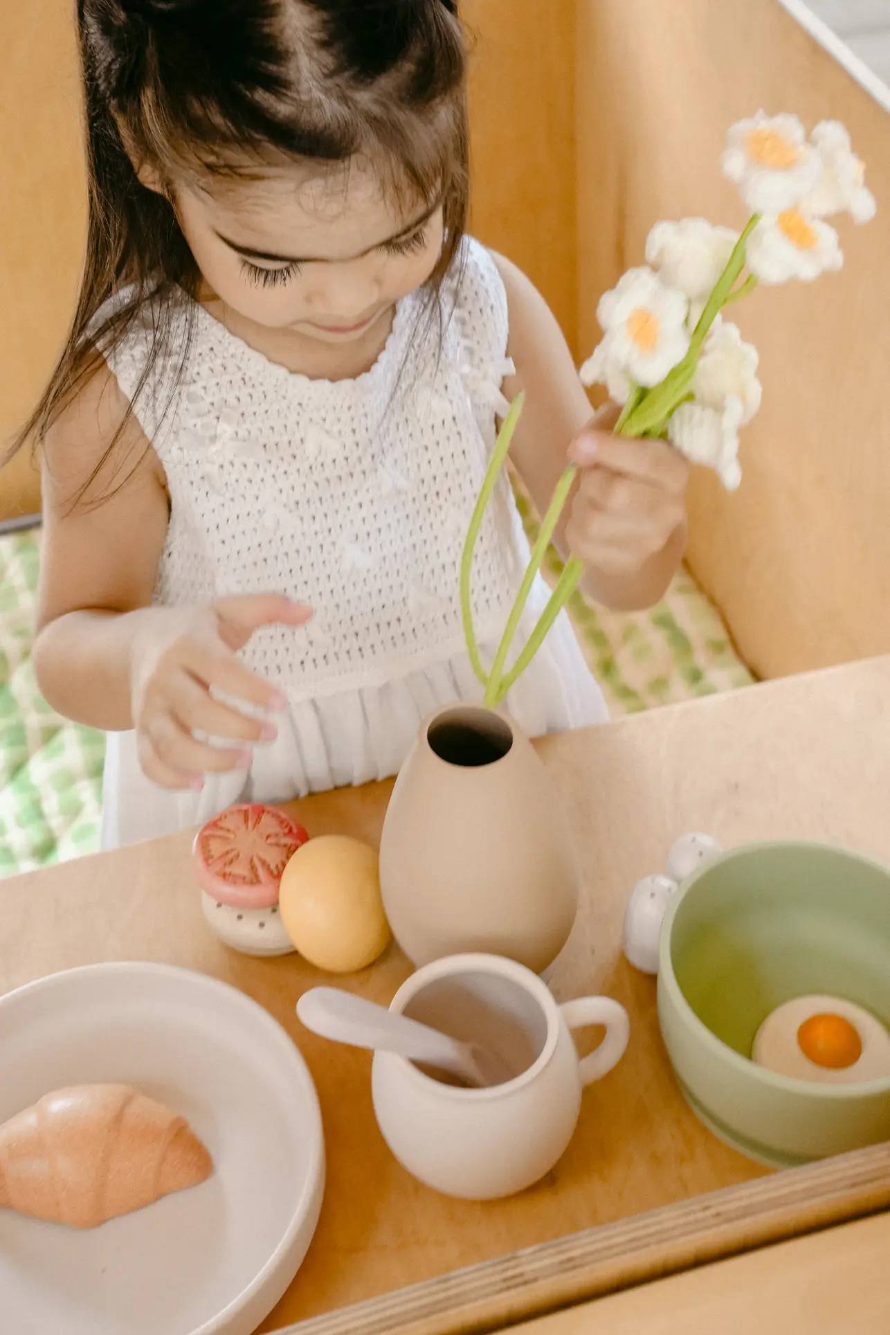A young girl in a white dress arranges white flowers into the Soft Spot Soft Vase by Soft Spot on a wooden table set with toy food, such as a croissant, an egg in a bowl, a cup, and a tomato.