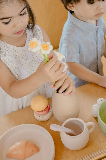 Two young children play at a wooden table with toy food, the Soft Spot Soft Vase by Soft Spot filled with fabric flowers, a cup with a spoon, a croissant on a plate, and other pretend dishes as they engage in imaginative play.