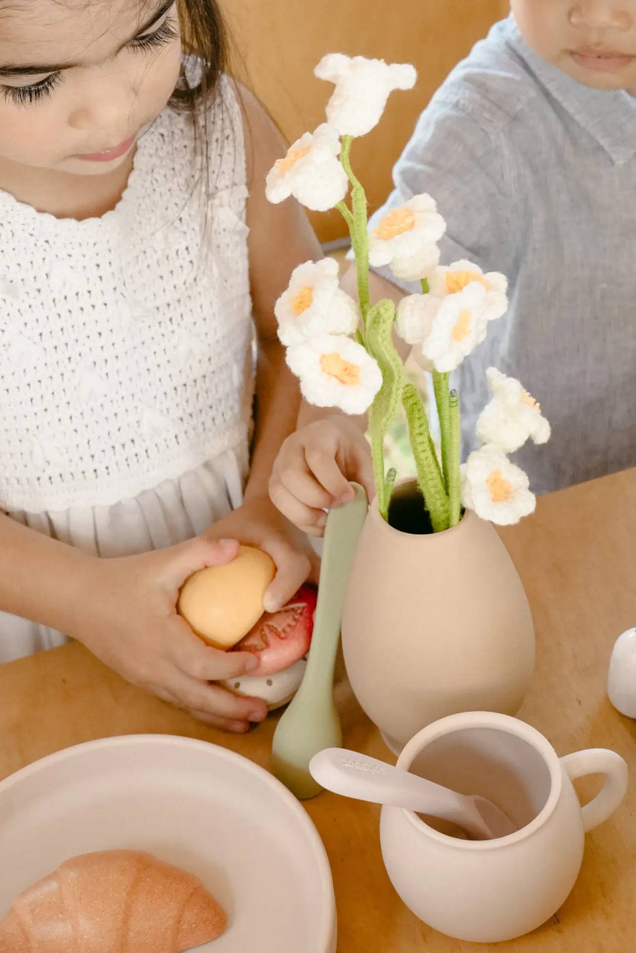 A young child in a white crochet dress holds toy food next to the Soft Spot Soft Vase by Soft Spot, filled with white and yellow felt flowers, on a wooden table set with toy dishes. Another child in a blue shirt is partially visible.