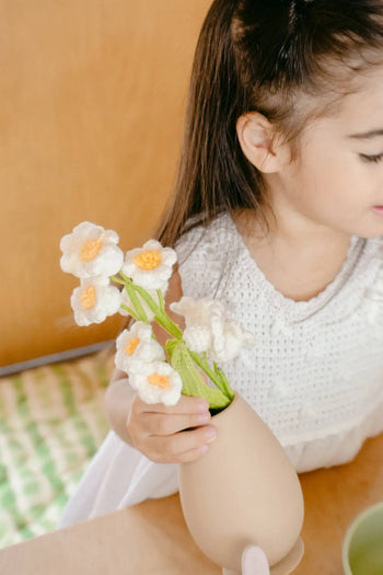 A young girl with long dark hair, wearing a white dress, sits at a table holding the Soft Spot Soft Vase by Soft Spot, filled with white and yellow crocheted flowers. She is partly turned away from the camera.