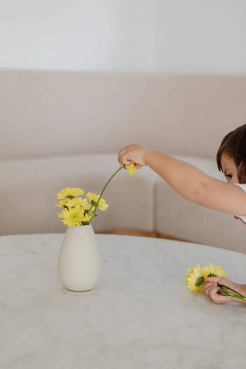 A child places a yellow flower into the Soft Spot Soft Vase by Soft Spot, an unbreakable and stylish vase with several blooms on a round, light-toned table. Behind it are a beige sofa and a plain wall.