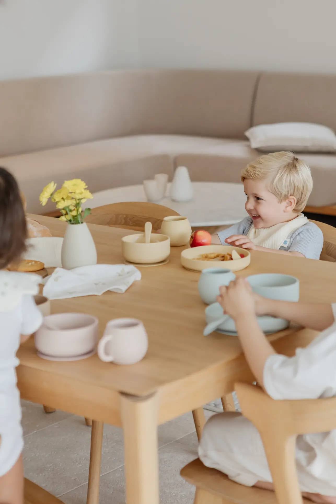 Three young children sit around a wooden table with pastel bowls and cups, enjoying a meal. At the center is the Soft Spot Soft Vase by Soft Spot, holding yellow flowers. A beige sofa is visible in the background.