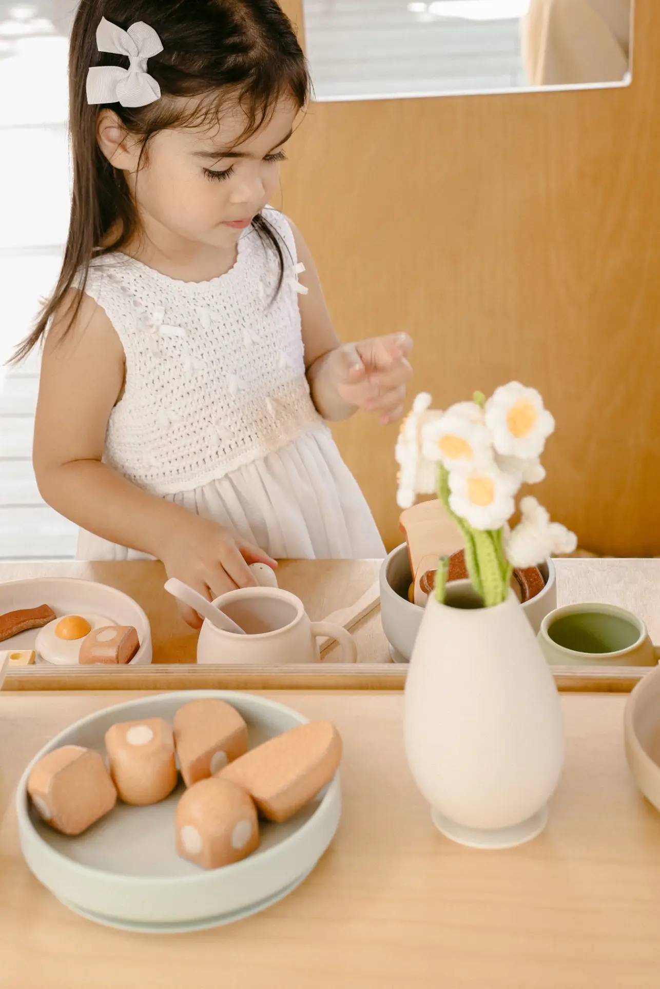 A young girl in a white dress plays with toy food and tea sets on a wooden table. In the foreground is the Soft Spot Soft Vase by Soft Spot, filled with felt flowers. The scene is bright and softly lit.