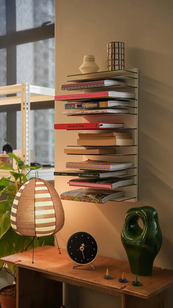 The Spectrum Paperback Wallsystem 30x60 by Spectrum displays vibrant books and notebooks above a wooden desk with a striped lamp, black clock, and green abstract sculpture. Minimalist décor is enhanced by plants and natural light from the window.