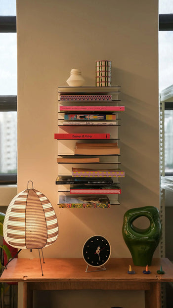 The Spectrum Paperback Wallsystem 30x60 by Spectrum is mounted on a beige wall, displaying colorful books and decor. Beneath it are a striped paper lantern, green ceramic sculpture, and black clock. Sunlight enhances the minimalist aesthetic.