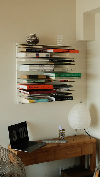 A minimalist workspace includes a wooden desk with an open laptop and a paper lantern lamp. Above, the Spectrum Paperback Wallsystem 60x60 by Spectrum displays books and vases against a neutral wall.