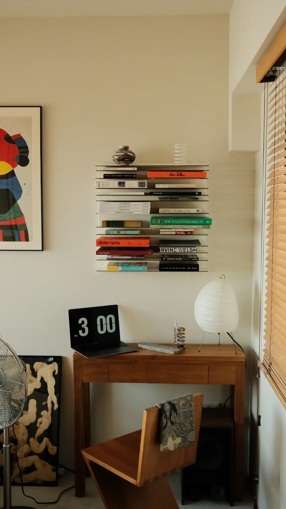 A minimalist workspace features a wooden desk, modern chair, digital clock at 3:00, paper lantern, Spectrum Paperback Wallsystem 60x60 by Spectrum stacked with books, and abstract art on a cream-colored wall.