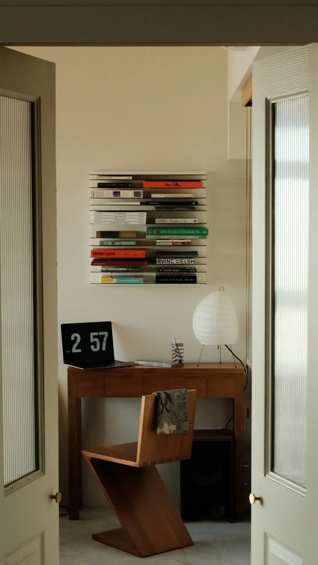 A small, minimalist workspace features a wooden desk, chair, digital clock at 2:57, a modern white lamp, and neatly stacked books on the Spectrum Paperback Wallsystem 60x60 by Spectrum. Light streams through open double doors.