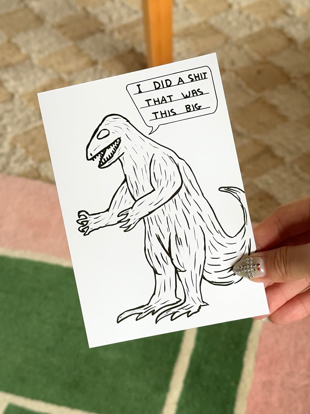 A hand holds the Stacked Store's Dinosaur Shit Gift Card (Physical), featuring a cartoon dinosaur with a cheeky speech bubble. Set on a green and pink rug, it's a quirky holiday gift—ideal for anyone with a sense of humor.
