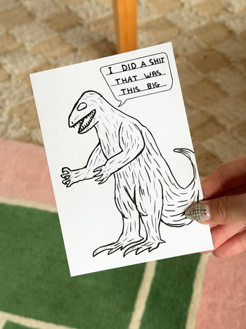 A hand holds the Stacked Store's Dinosaur Shit Gift Card (Physical), featuring a cartoon dinosaur with a cheeky speech bubble. Set on a green and pink rug, it's a quirky holiday gift—ideal for anyone with a sense of humor.