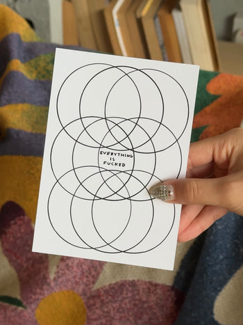 A hand with decorated nails holds the Stacked Store Everything Is Fucked Gift Card (Physical) by David Shrigley, displaying overlapping black circles and bold text—perfect for quirky holiday gifting amidst colorful books and fabrics.