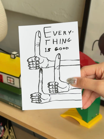 A hand holds the Stacked Store’s Everything Is Good Gift Card (Physical), featuring illustrated hands forming E, V, and L in sign language with “EVERYTHING IS GOOD” in handwritten text—perfect for holiday gifting. Toy house decor shown in background.