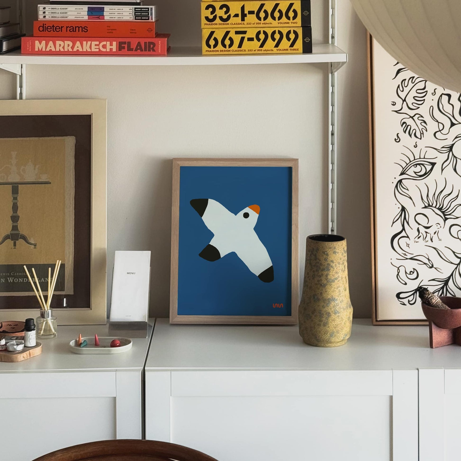 The Stacked Store Exclusive Bernard de Blur Poster (30x40) is displayed on a white cabinet with vases, artwork, and books in a modern living space. It features a stylized blue bird in flight and adds an artistic touch to the room.