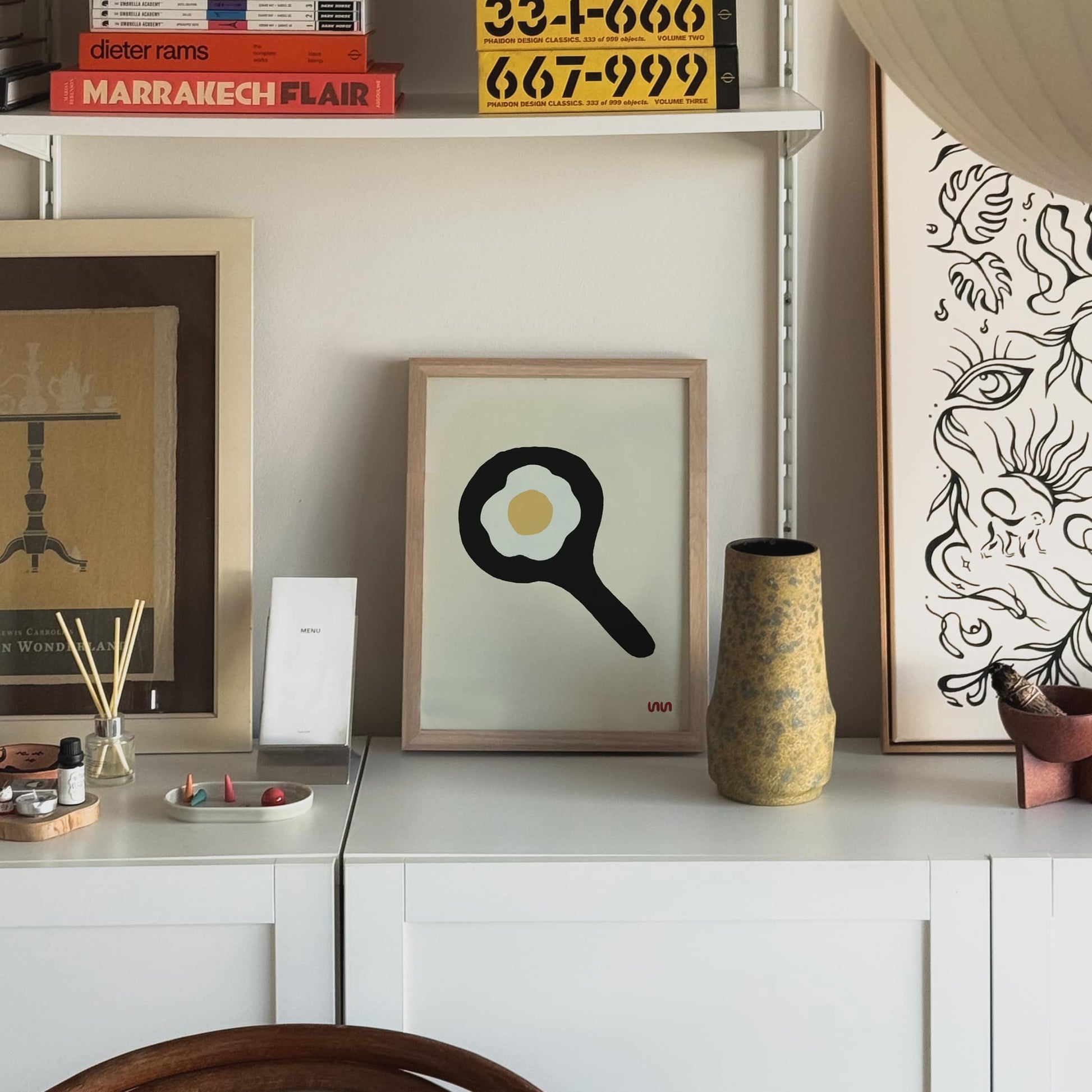 The Stacked Store Exclusive Big Eggo Poster (30x40) is displayed on a white cabinet, surrounded by unique decor, posters, books, and a vase in a cozy, modern room.