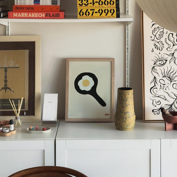 The Stacked Store Exclusive Big Eggo Poster (30x40) is displayed on a white cabinet, surrounded by unique decor, posters, books, and a vase in a cozy, modern room.