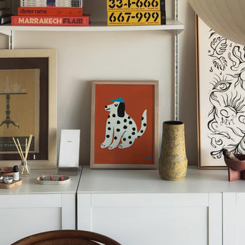 The Stacked Store Exclusive Xavier The Dog Poster (30x40) by Stacked Store, featuring a Dalmatian in a blue cap, is displayed on a white cabinet amid art, books, and decor in a modern room.