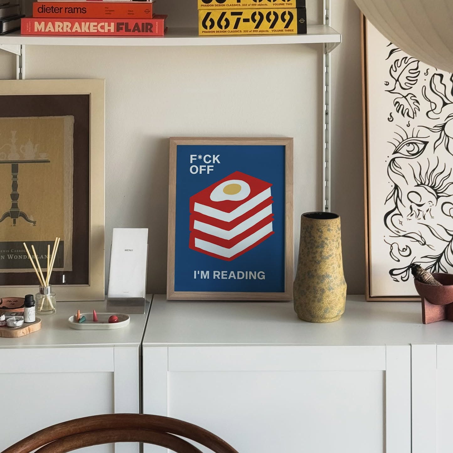 The Stacked Store Fuck Off Im Reading - The Great Yolksby Poster (30x40) sits on a white cabinet, featuring book stacks and a fried egg. Surrounded by art, shelves, and decor, it adds playful charm to this cozy modern room.
