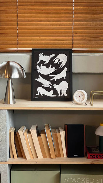 The Stacked Store Jaeyyelle - Cat Poster (30x40) by Stacked Store, featuring white cat silhouettes, is framed and displayed on a shelf next to a silver lamp, white clock, books, and a speaker. Wooden blinds hang in the background.