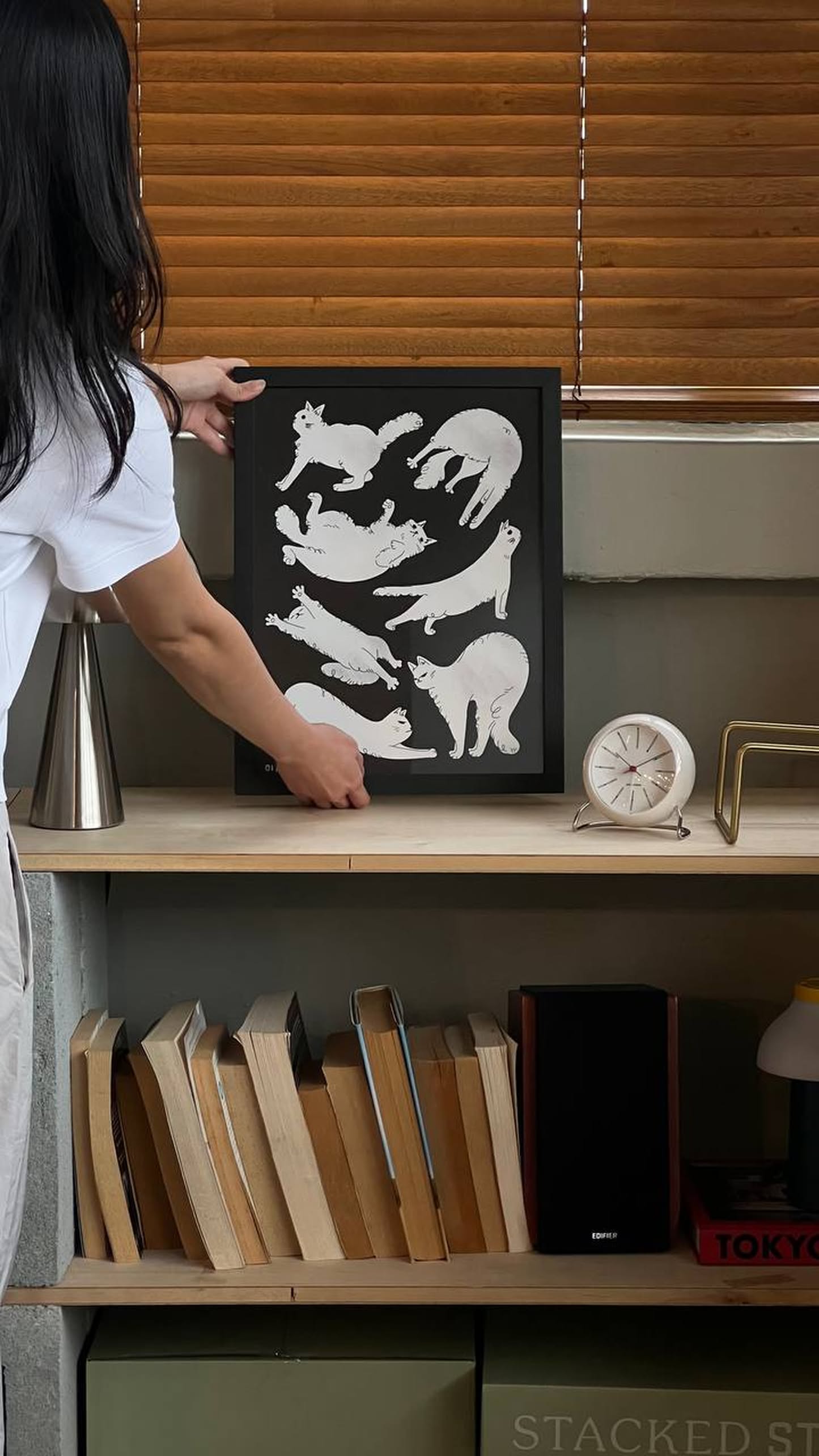 A person places the Stacked Store Jaeyyelle - Cat Poster (30x40) on a shelf with books, a clock, a speaker, and other items. Wooden blinds cover the window above.