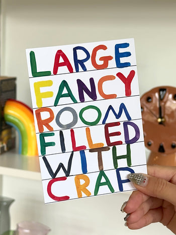 A hand holds a vibrant sign reading "LARGE FANCY ROOM FILLED WITH CRAP." Shelves adorned with decor add charm—ideal for gifting alongside the Large Fancy Room Gift Card (Physical) from Stacked Store.