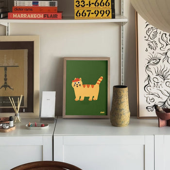 The Stacked Store Exclusive Scaredy Cat Poster (30x40) is displayed on a white cabinet amid decor, art prints, and books, making it a charming feature for cat lovers from Stacked Store.