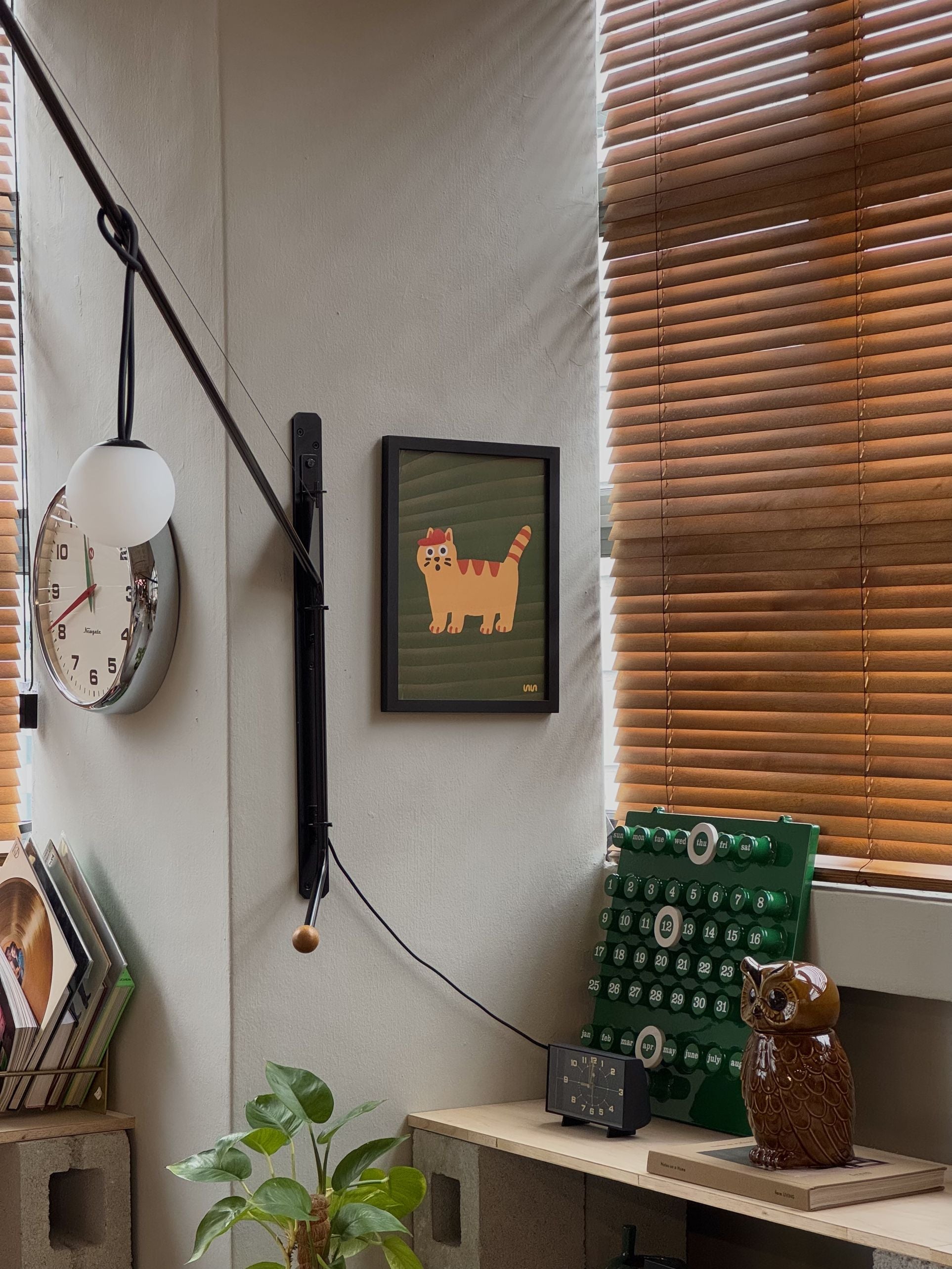 A cozy corner displays a Stacked Store Exclusive Scaredy Cat Poster (30x40) on the wall, a round clock, vintage green typewriter, owl figurine, wooden blinds, and books. Sunlight brings warmth to the inviting space.