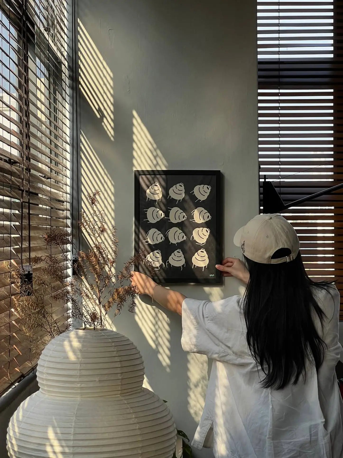 A person in a white shirt and beige cap hangs the Stacked Store Trip Poster by Stacked Store on a sunlit wall, with striped shadows from window blinds. Decorative branches and an Akari 24N lamp add minimalist elegance to the scene.