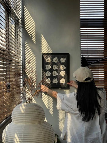 A person in a white shirt and beige cap hangs the Stacked Store Trip Poster by Stacked Store on a sunlit wall, with striped shadows from window blinds. Decorative branches and an Akari 24N lamp add minimalist elegance to the scene.