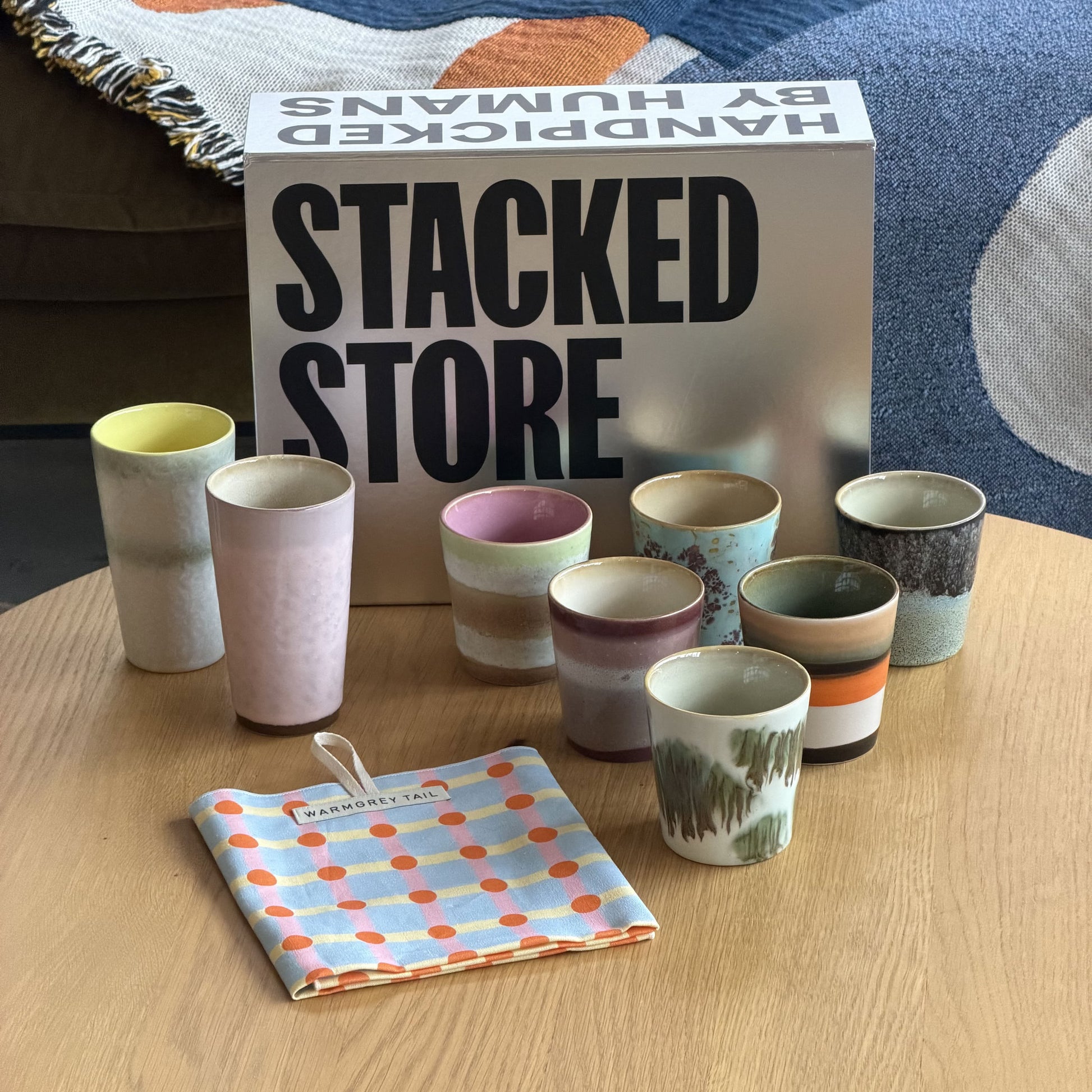 A set of vibrant HKLIVING 70s Ceramics cups and a small pitcher sit on a wooden table with a striped napkin. Behind them is the Stacked Store The Coffee Lover Gift Box by Stacked Store, with patterned fabric in the background.