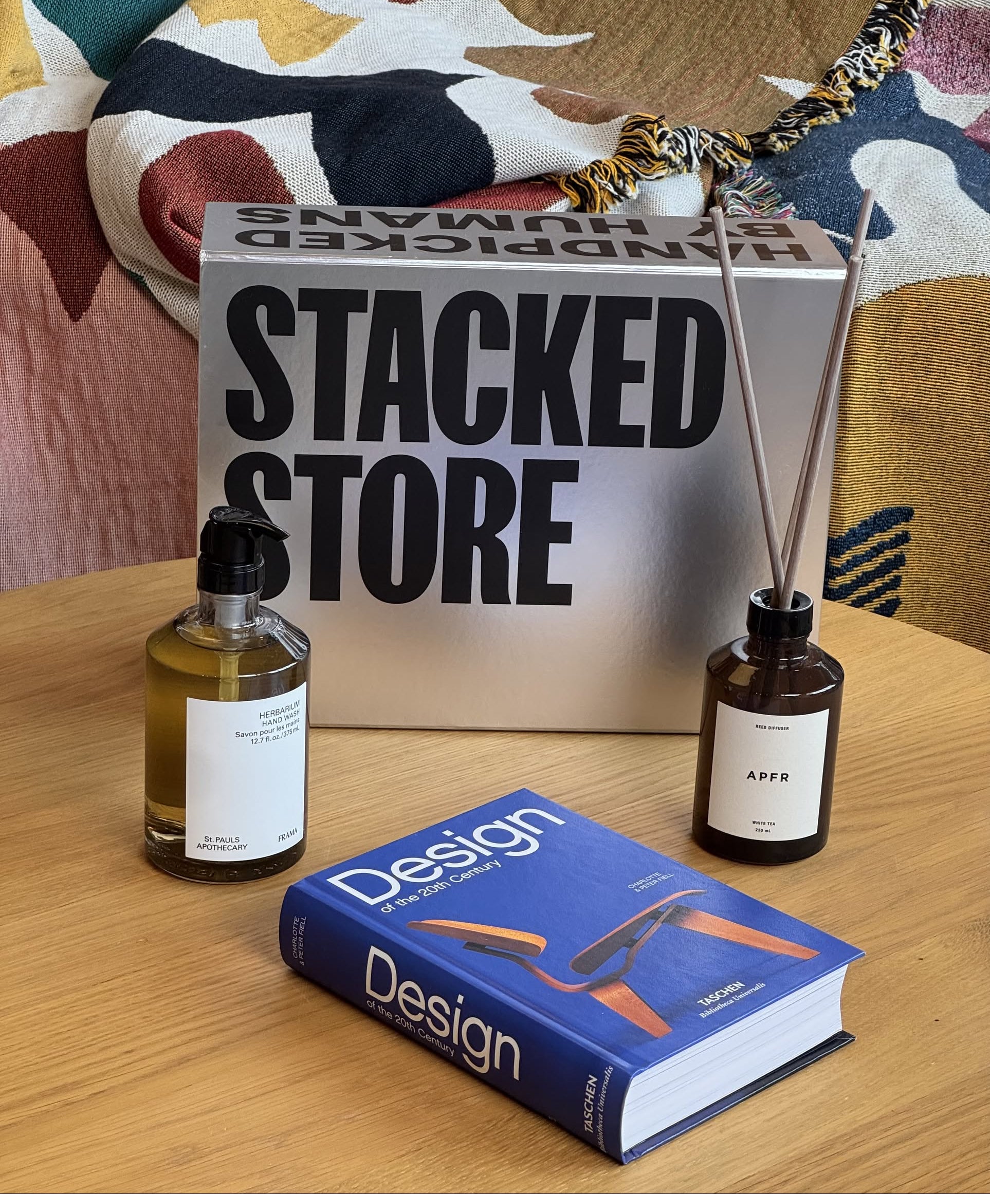 A tabletop displays Stacked Store’s The Design Enthusiast Gift Box—featuring a design book, labeled glass bottle, APFR Reed Diffuser, and a reflective STACKED STORE box—set on colorful patterned fabric.