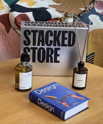 A tabletop displays Stacked Store’s The Design Enthusiast Gift Box—featuring a design book, labeled glass bottle, APFR Reed Diffuser, and a reflective STACKED STORE box—set on colorful patterned fabric.