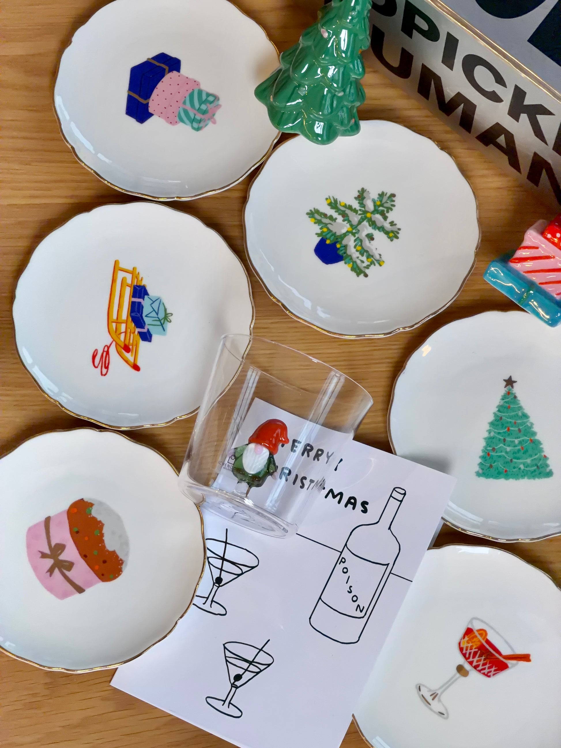 The Festive Holiday Gift Box by Stacked Store includes seven small Christmas plates with festive designs and a Merry Christmas card featuring a wine bottle and two figures—ideal for gifting or same-day delivery, beautifully arranged on a wooden surface.
