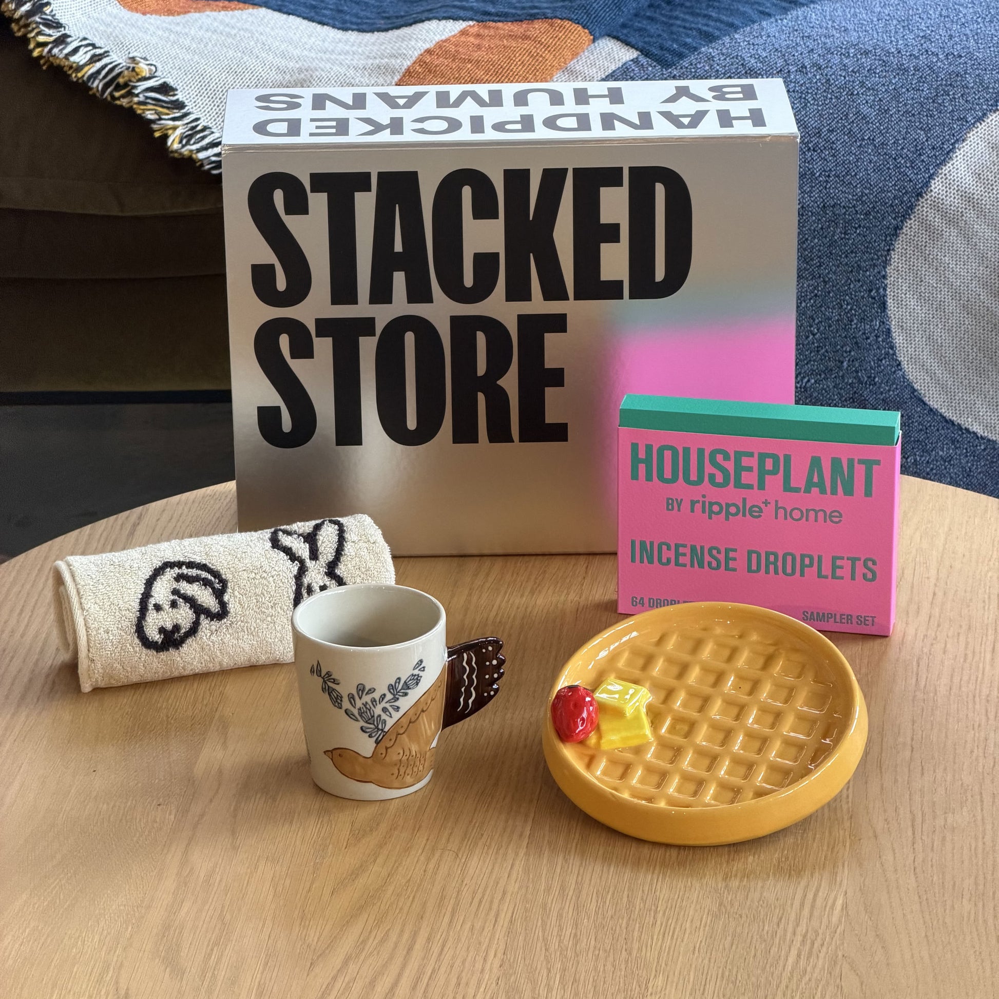 A wooden table displays the Stacked Store The Homebody Gift Box, featuring a towel with faces, orange ceramic pancakes with blueberries, a face cup, colorful incense cones, and branded packaging—all from Stacked Store. Enjoy same-day delivery!.