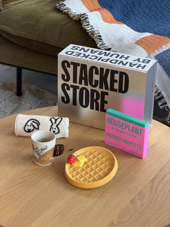 A cozy setup showcases two Stacked Store The Homebody Gift Boxes, a cup with faces, a rolled towel, pancake-shaped coasters, and a pink incense droplet box on a patterned blanket—perfect for same-day delivery.