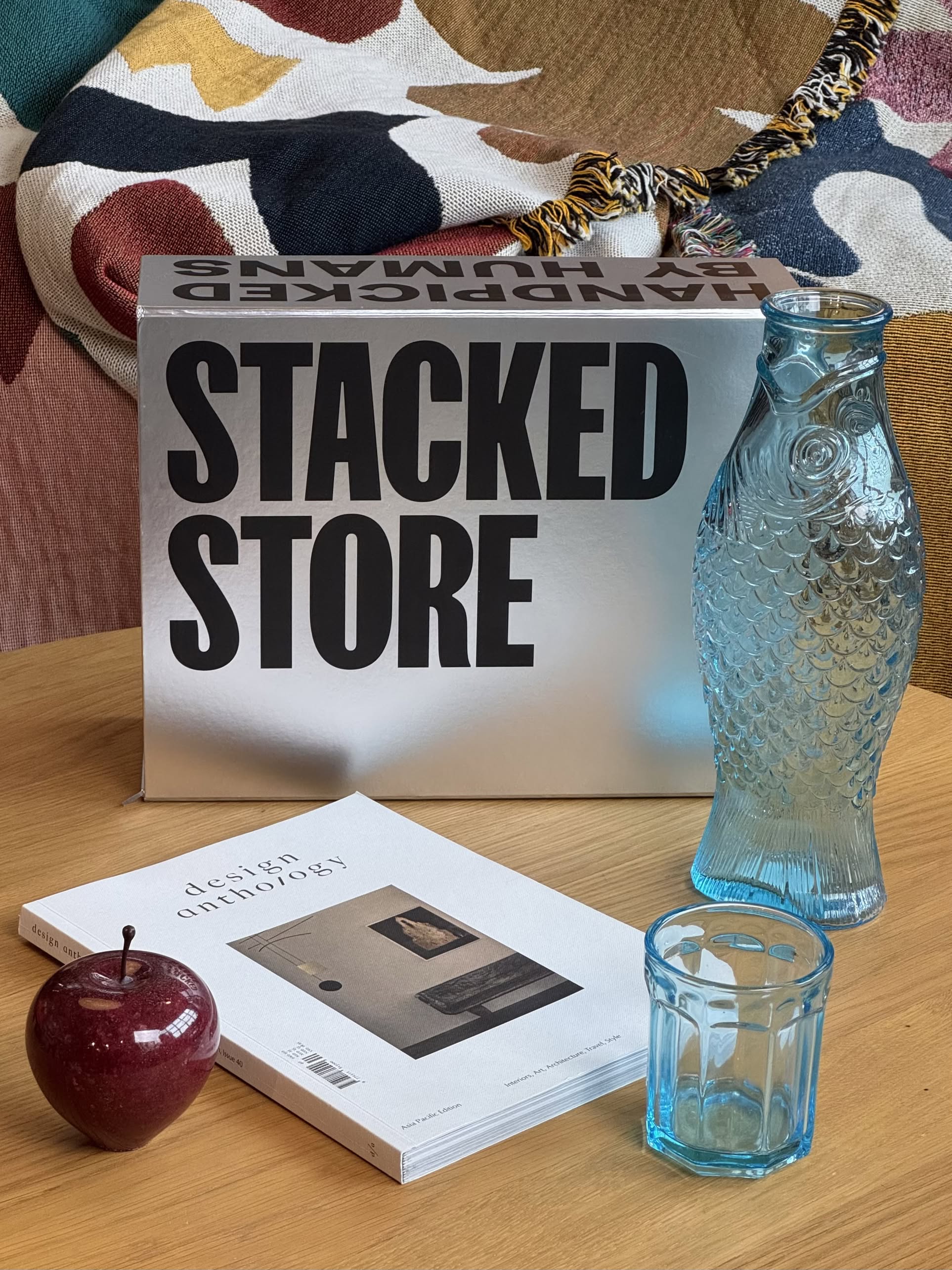 A tabletop displays the Stacked Store The Minimalists Gift Box, a blue fish-shaped glass bottle, a blue glass cup, a magazine, and a red apple. A patterned blanket is draped in the background—a stylish gift ready for same-day delivery.