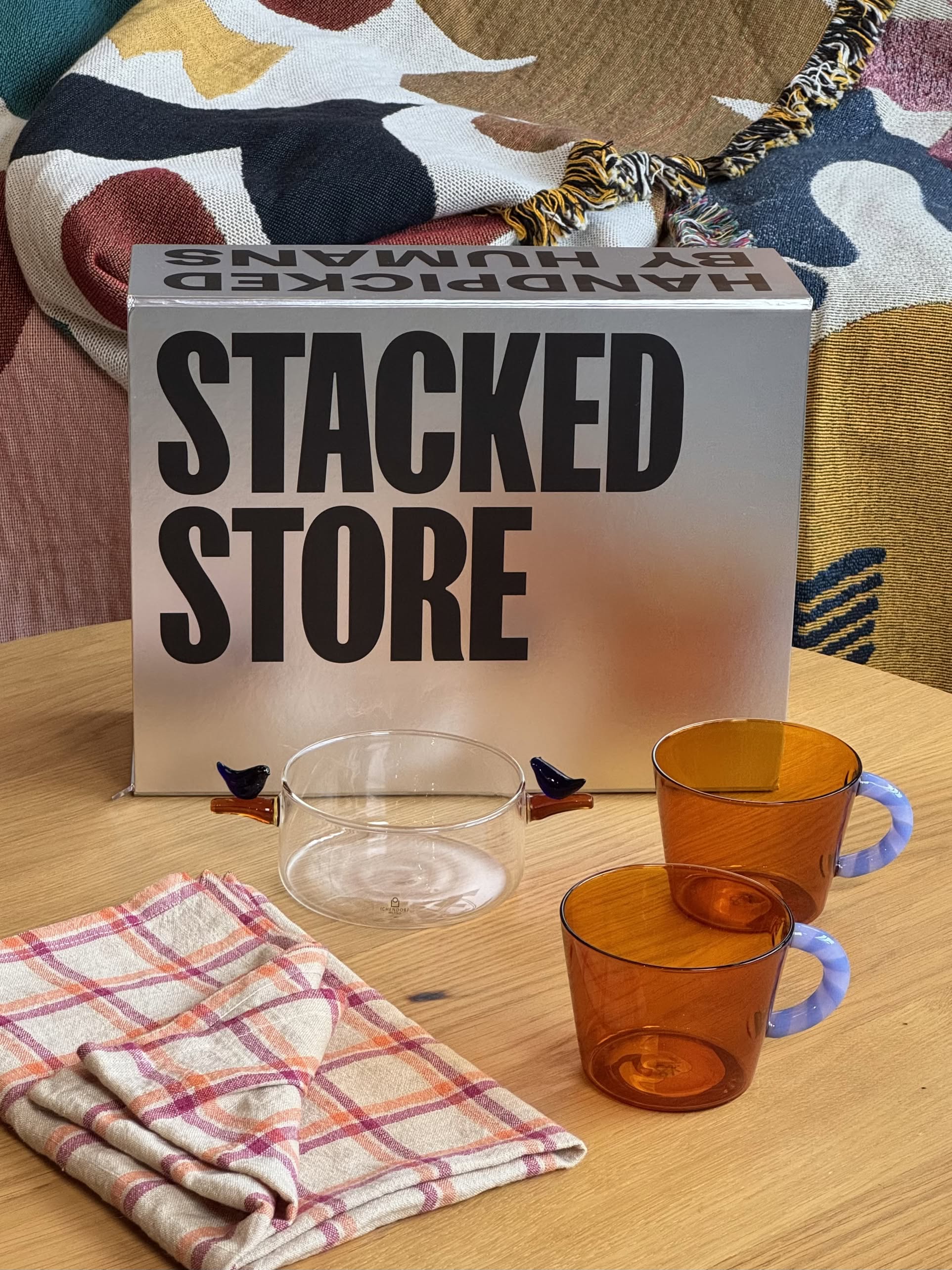 A display on a wooden table features the Stacked Store The Modern Host Gift Box, amber glasses with lavender handles, a clear glass cup with blue and brown handles, and a colorful patterned blanket in the background.