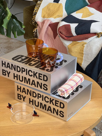 Two amber glasses and a rolled plaid napkin from the Stacked Store The Modern Host Gift Box rest atop silver boxes, with a handcrafted glass bowl nearby and a colorful blanket on a green chair in the background.