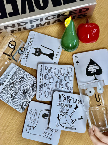 A rainbow ornament, unique gift box, two animal-themed glasses, and two ceramic fish are displayed on a wooden table before a silver Stacked Store The People Who Have Everything Gift Box, with a patterned blanket in the background.
