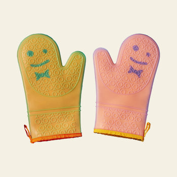 Staff The Oven Mitts, Set of 2 by Staff includes colorful silicone mitts with smiley faces—one yellow with green and blue accents, one pink with purple. Both are temperature-resistant, textured for grip, and feature hanging loops for convenience.