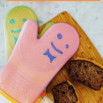 Staff The Oven Mitts, Set of 2 by Staff feature playful, colorful designs with smiling faces and are shown on a wooden board beside chocolate chip bread, showcasing their temperature resistance and cheerful appeal.