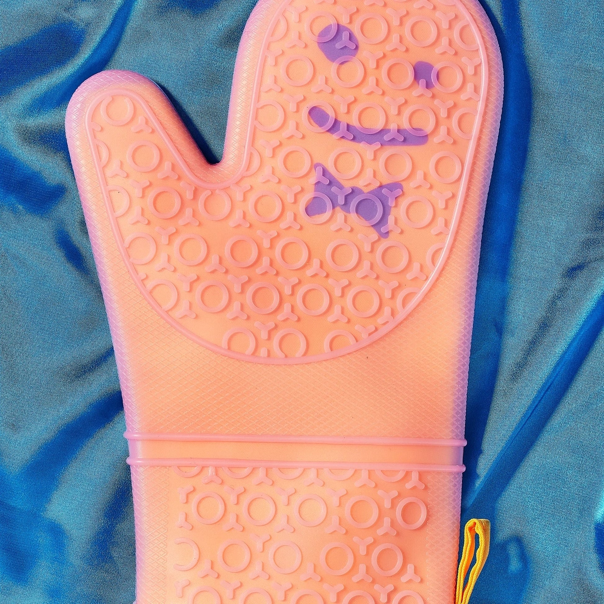 Staff The Oven Mitts, Set of 2 by Staff are orange silicone mitts featuring a textured raised circular pattern and smiling face, known for great temperature resistance, shown on a shiny blue fabric background.