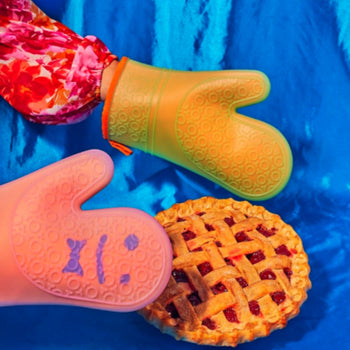Someone wearing colorful, floral sleeves and using Staff The Oven Mitts, Set of 2 by Staff—renowned for heat resistance—holds a freshly baked lattice pie on a vibrant blue fabric background.